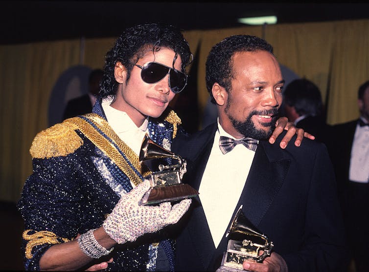 A young man wearing sunglasses and a bejeweled outfit poses with a gold trophy of a small record player next to a goateed man also holding the same trophy.